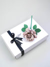 Grey tin paper rose lying diagonally on top of a luxury white gift box that has a black satin ribbon tied in a bow with a Miss Poppins and Me gift tag attached