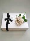 Shimmery pearl paper rose lying diagonally on top of a luxury white gift box that has a black satin ribbon tied in a bow with a Miss Poppins and Me gift tag attached