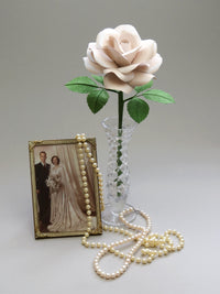 Pearl paper rose with six ivy green leaves standing in a slender glass vase to the right of a vintage gold photo frame with a sepia photo of a happy vintage bride and groom, with a long stranded pearl necklace delicately laid over the right corner of the frame and sprawled across the foreground of the photo