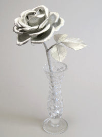 Silver crepe paper rose with three silver leaves standing in a narrow glass vase against a light grey backdrop