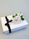 White linen grain paper rose lying on top of a luxury white gift box that has a black satin ribbon tied in a bow with a Miss Poppins and Me gift tag attached